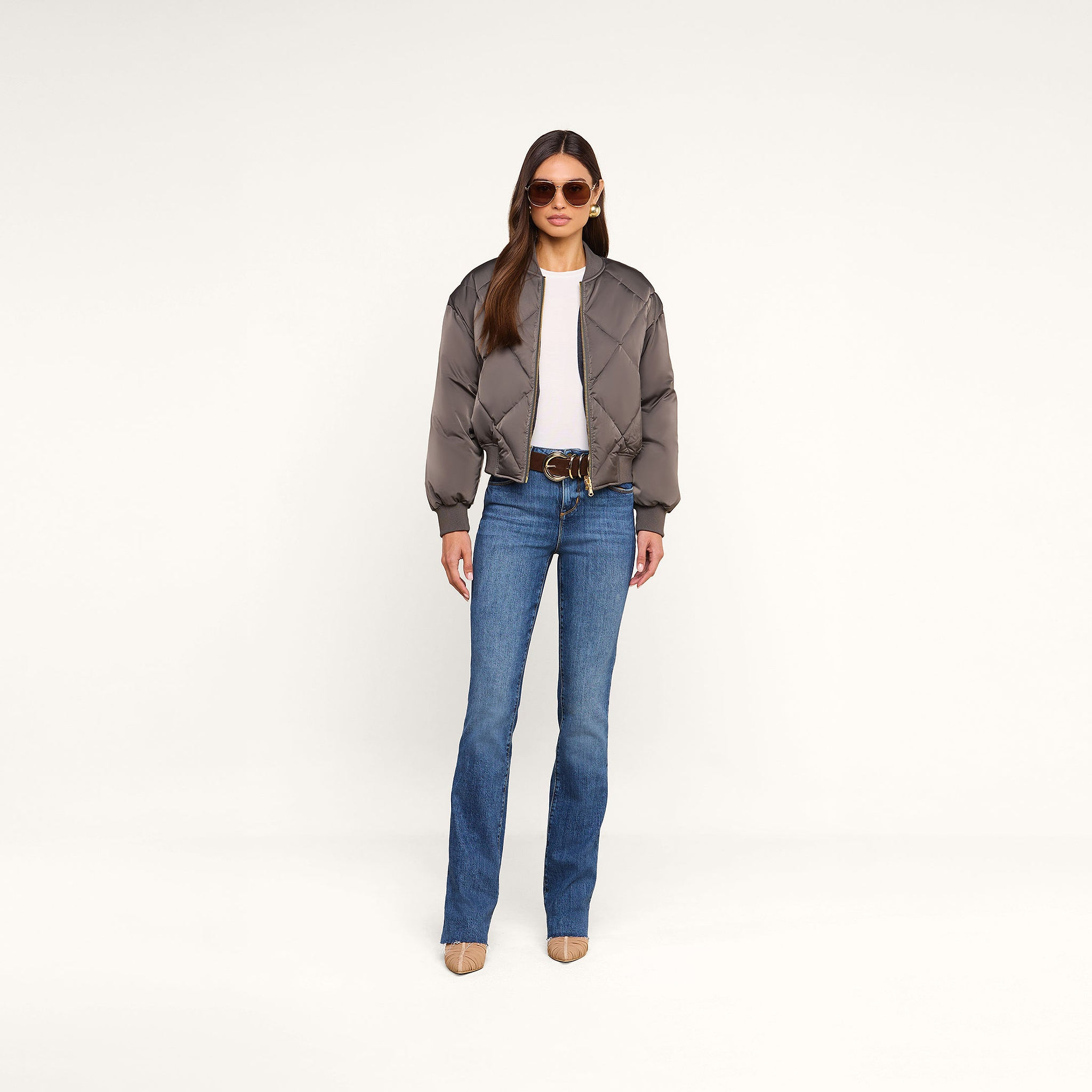 A woman wears sunglasses, a white top, blue flared jeans, beige heels, and the Violeta Reversible Puffer—a gray quilted satin bomber jacket—as she stands against a beige background facing forward with her hands by her sides.