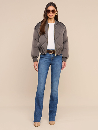A woman wears sunglasses, a white top, blue flared jeans, beige heels, and the Violeta Reversible Puffer—a gray quilted satin bomber jacket—as she stands against a beige background facing forward with her hands by her sides.