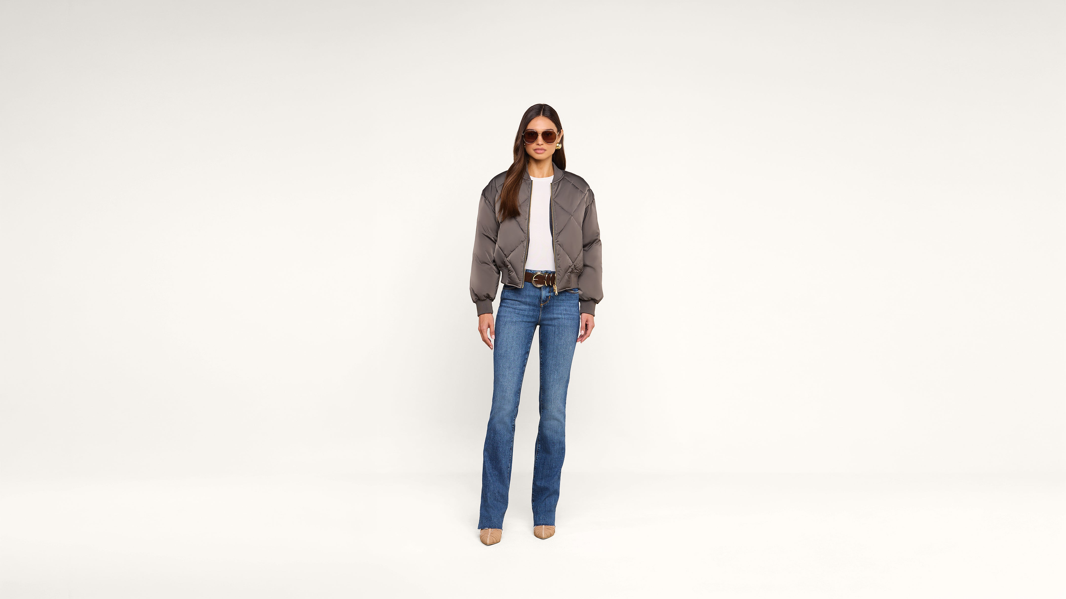 A woman stands against a plain white background wearing sunglasses, a gray jacket, white shirt, blue jeans, and beige shoes.