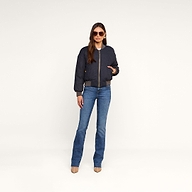 A woman wearing sunglasses, the Violeta Reversible Puffer, blue jeans, and beige heels stands against a plain beige background.