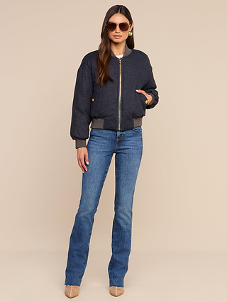 A woman wearing sunglasses, the Violeta Reversible Puffer, blue jeans, and beige heels stands against a plain beige background.