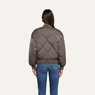 A person stands facing away, wearing the Violeta Reversible Puffer in gray with ribbed cuffs and blue jeans, against a plain beige background.