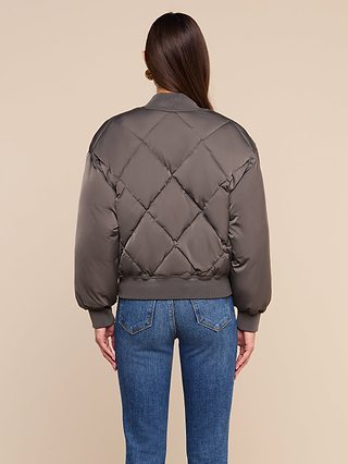 A person stands facing away, wearing the Violeta Reversible Puffer in gray with ribbed cuffs and blue jeans, against a plain beige background.