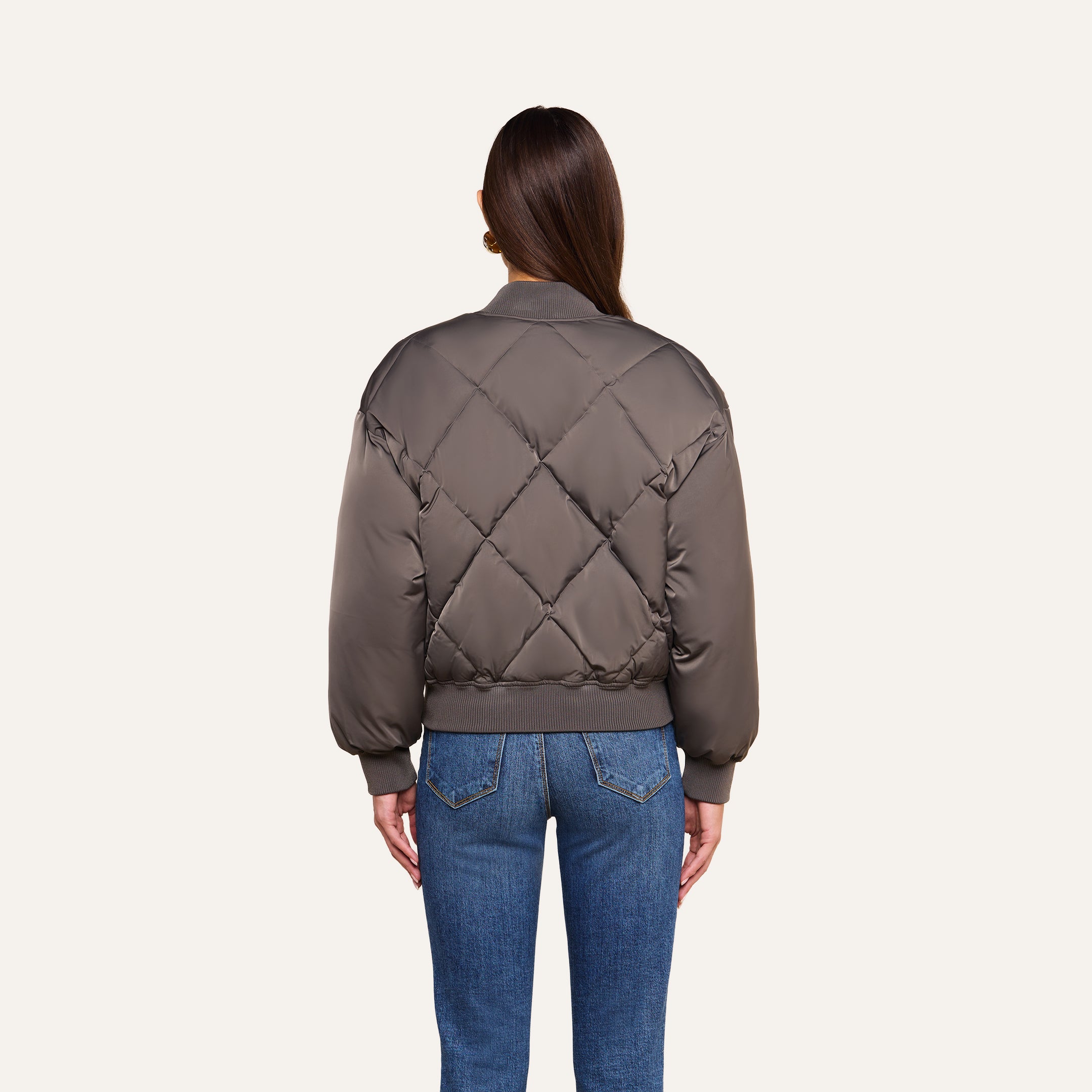 A person stands facing away, wearing the Violeta Reversible Puffer in gray with ribbed cuffs and blue jeans, against a plain beige background.