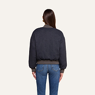 A woman with long brown hair, seen from behind, wears the Violeta Reversible Puffer with ribbed cuffs and waistband, paired with blue jeans, standing against a plain beige background.