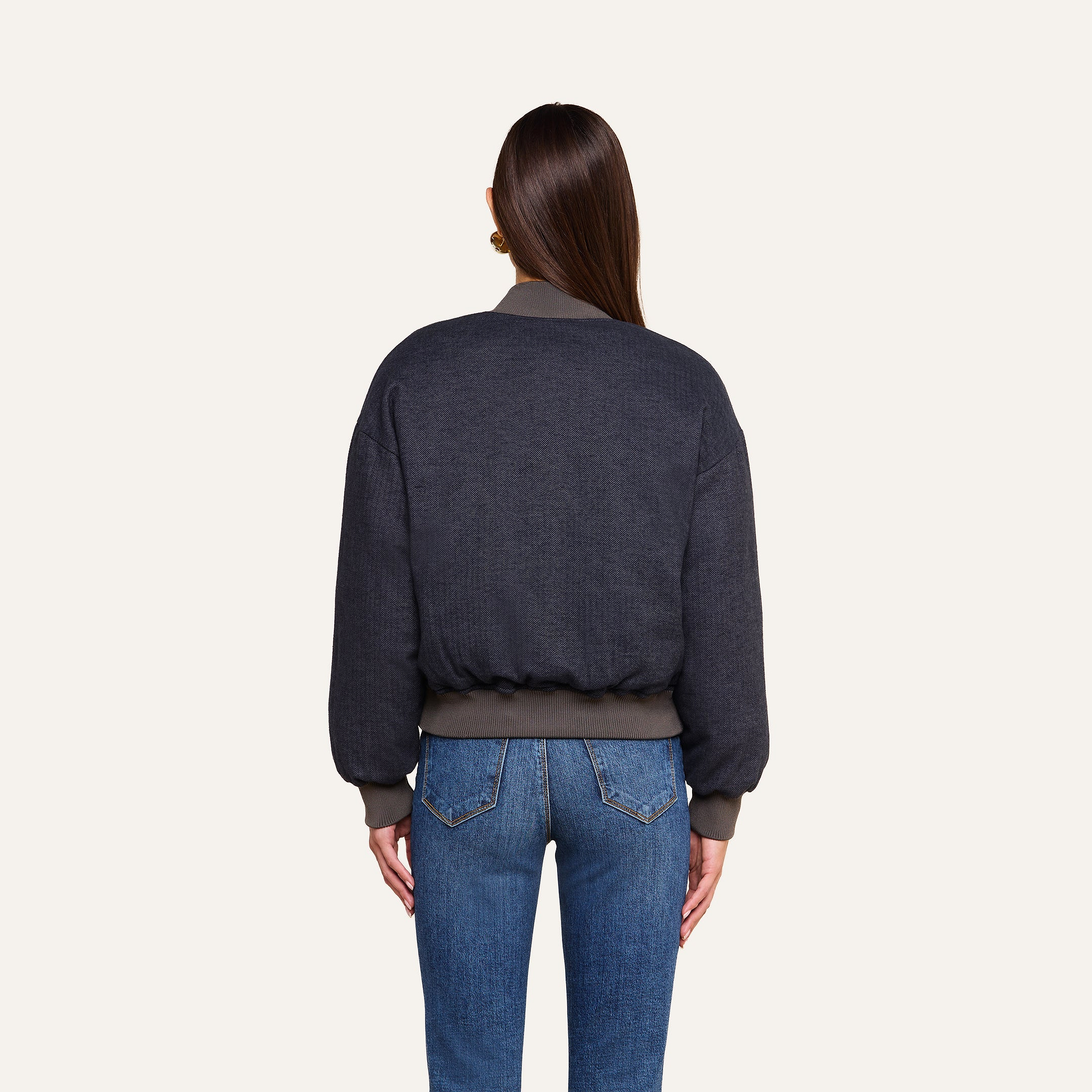 A woman with long brown hair, seen from behind, wears the Violeta Reversible Puffer with ribbed cuffs and waistband, paired with blue jeans, standing against a plain beige background.
