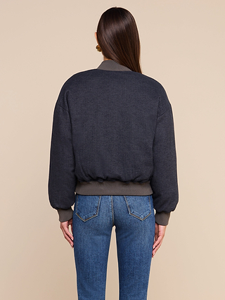 A woman with long brown hair, seen from behind, wears the Violeta Reversible Puffer with ribbed cuffs and waistband, paired with blue jeans, standing against a plain beige background.