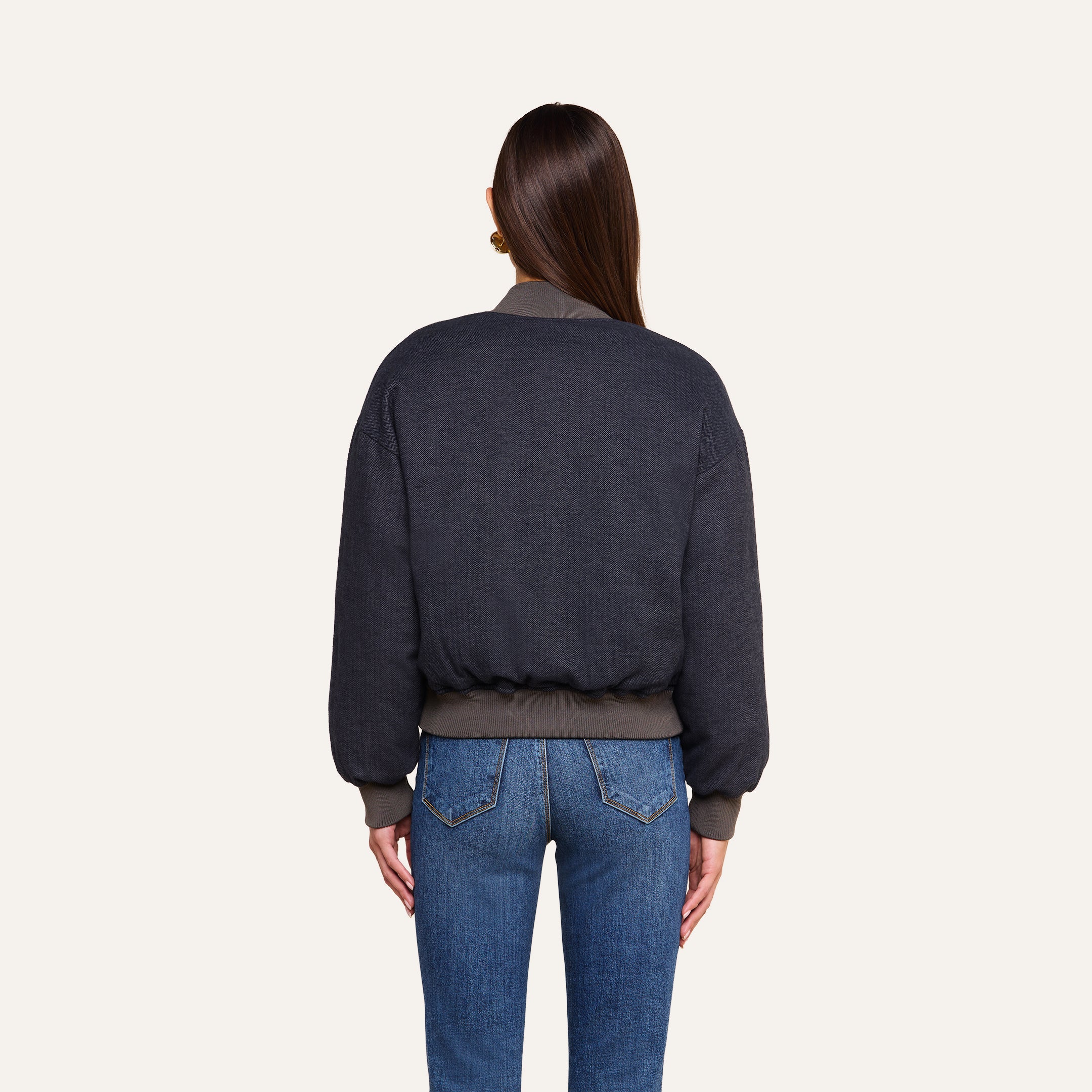A woman with long brown hair, seen from behind, wears the Violeta Reversible Puffer with ribbed cuffs and waistband, paired with blue jeans, standing against a plain beige background.