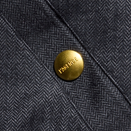 Close-up of a gray herringbone weave fabric from the Violeta Reversible Puffer, featuring a shiny gold button engraved with the word FRANCE. The button is attached to a stitched seam on the garment, recalling details found on classic reversible bomber jackets.