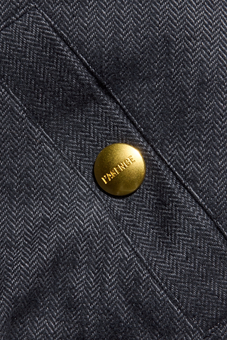 Close-up of a gray herringbone weave fabric from the Violeta Reversible Puffer, featuring a shiny gold button engraved with the word FRANCE. The button is attached to a stitched seam on the garment, recalling details found on classic reversible bomber jackets.