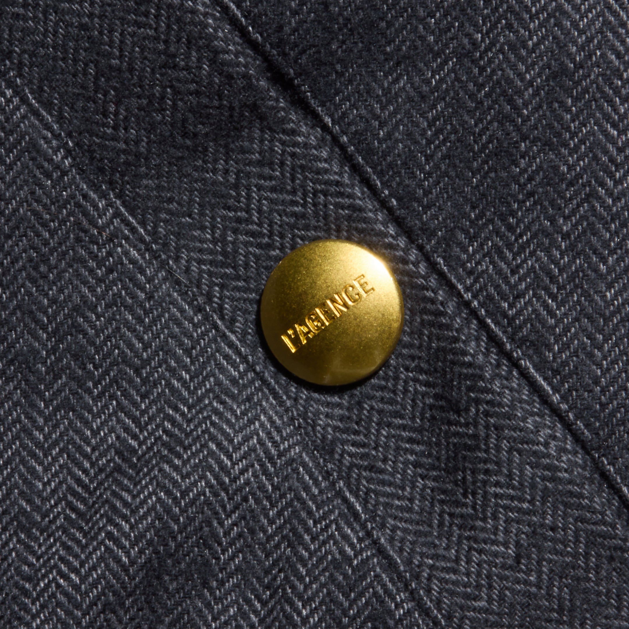 Close-up of a gray herringbone weave fabric from the Violeta Reversible Puffer, featuring a shiny gold button engraved with the word FRANCE. The button is attached to a stitched seam on the garment, recalling details found on classic reversible bomber jackets.