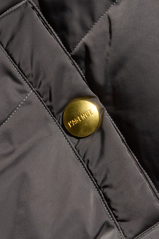 Close-up of a shiny gold button engraved with the word PATENCE on quilted black fabric, featured on the Violeta Reversible Puffer jacket.