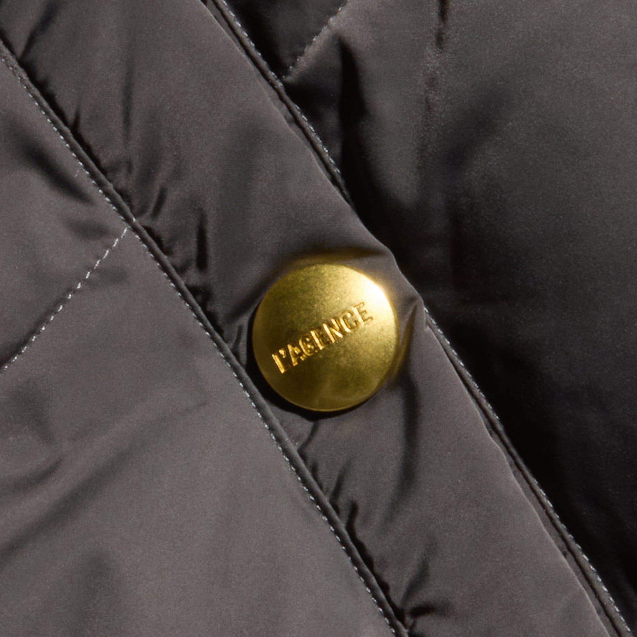 Close-up of a shiny gold button engraved with the word PATENCE on quilted black fabric, featured on the Violeta Reversible Puffer jacket.