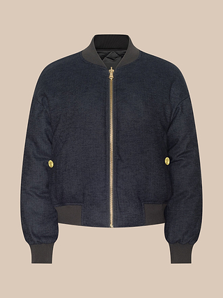 The Violeta Reversible Puffer is a navy blue bomber jacket featuring a gold zipper, gold buttons on the sides, and ribbed black collar, cuffs, and hem, showcased against a neutral beige background.