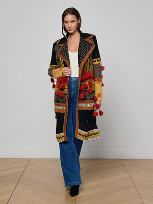 A woman stands indoors wearing the Kada Crochet Coat—a colorful patchwork long cardigan with red tassels—paired with a white top, blue wide-leg jeans, and black shoes on a wooden floor against a light background.