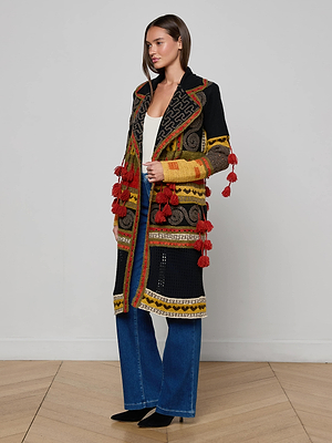 A woman with straight, shoulder-length hair stands indoors in the Kada Crochet Coat, a bohemian jacket featuring red tassels and intricate black, yellow, red, and brown designs, worn over a white top and blue jeans.