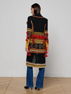A woman stands indoors on a light wooden floor, facing away, wearing the Kada Crochet Coat—a long bohemian cardigan with red tassels and yellow, orange, and black patterns—paired with blue jeans against a white wall.