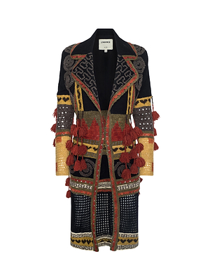 The Kada Crochet Coat is a knee-length, long-sleeve crochet coat with multicolored red, yellow, gold, black, and gray geometric patterns and textured tassels, shown on a white background.