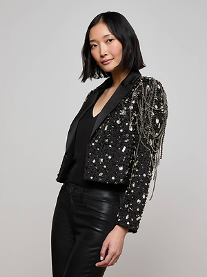 A woman with a chin-length black bob wears the Soma Embellished Blazer—detailed with hand-embellished crystals and silver chain fringe—over a black top and leather pants, standing against a plain light background.
