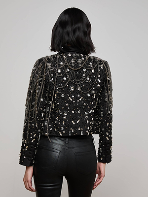 A person with shoulder-length dark hair, seen from behind, wears the Soma Embellished Blazer—black with a satin lapel, hand-embellished crystals, and hanging chains—paired with black pants. The background is plain and light-colored.