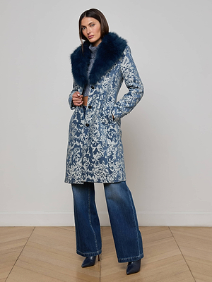 A woman poses against a light wall and wooden floor, wearing the L'AGENCE Laney Jacquard Coat with a dark blue faux fur collar, blue denim flared jeans, and navy pointed-toe shoes.