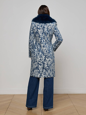 A person stands facing away, wearing the Laney Jacquard Coat by L'AGENCE with a dark blue faux fur collar, paired with wide-leg blue jeans and black heeled shoes on a wooden floor.