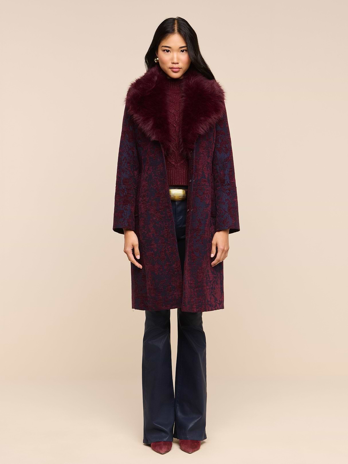 A woman stands against a white background, wearing the Laney Jacquard Coat—a long burgundy coat with a large faux fur collar—paired with navy trousers and burgundy pointed shoes.