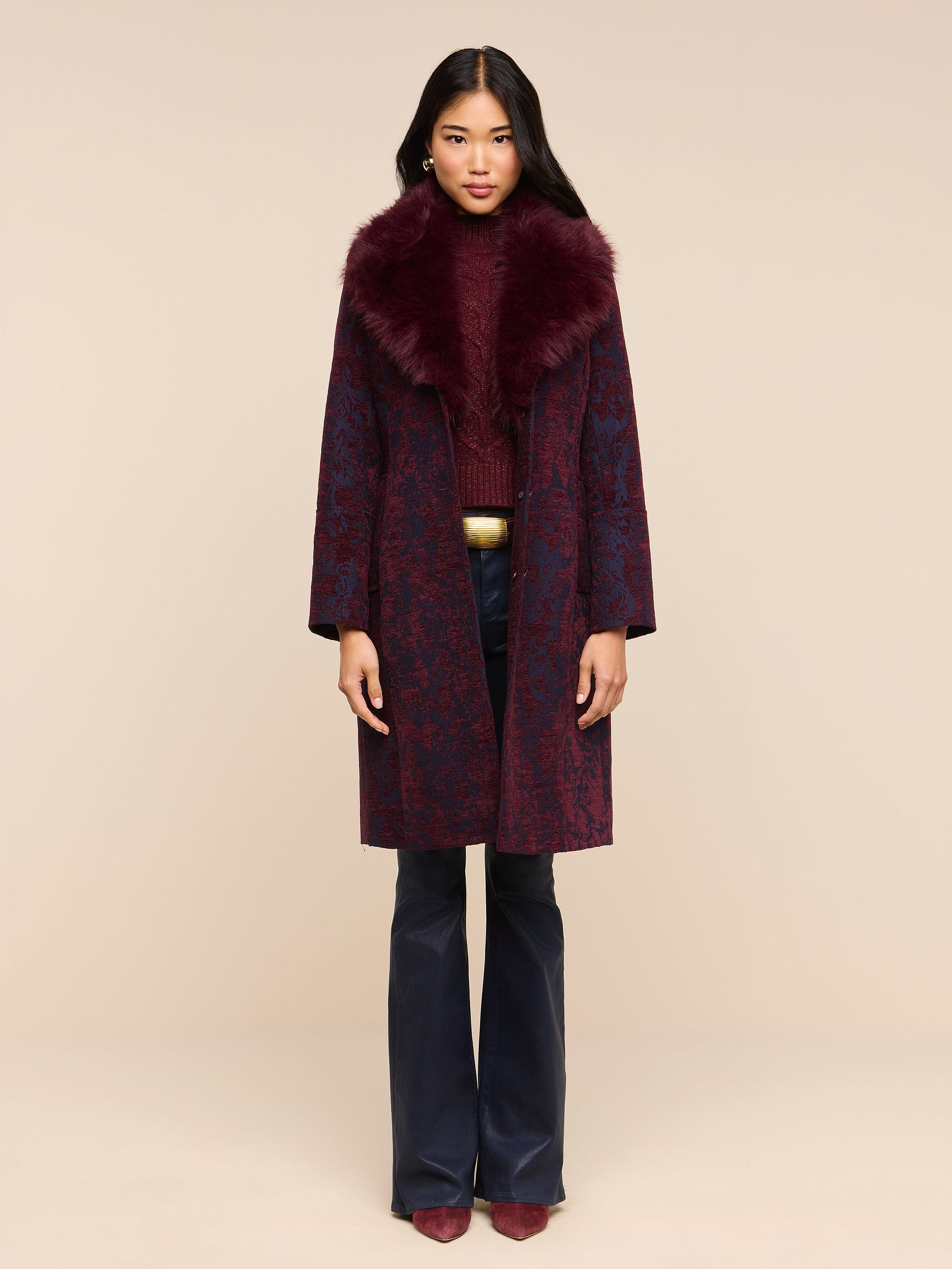 A model wears a long patterned coat with a dark burgundy faux fur collar over a matching sweater and dark Marty Coated Flare Jean.