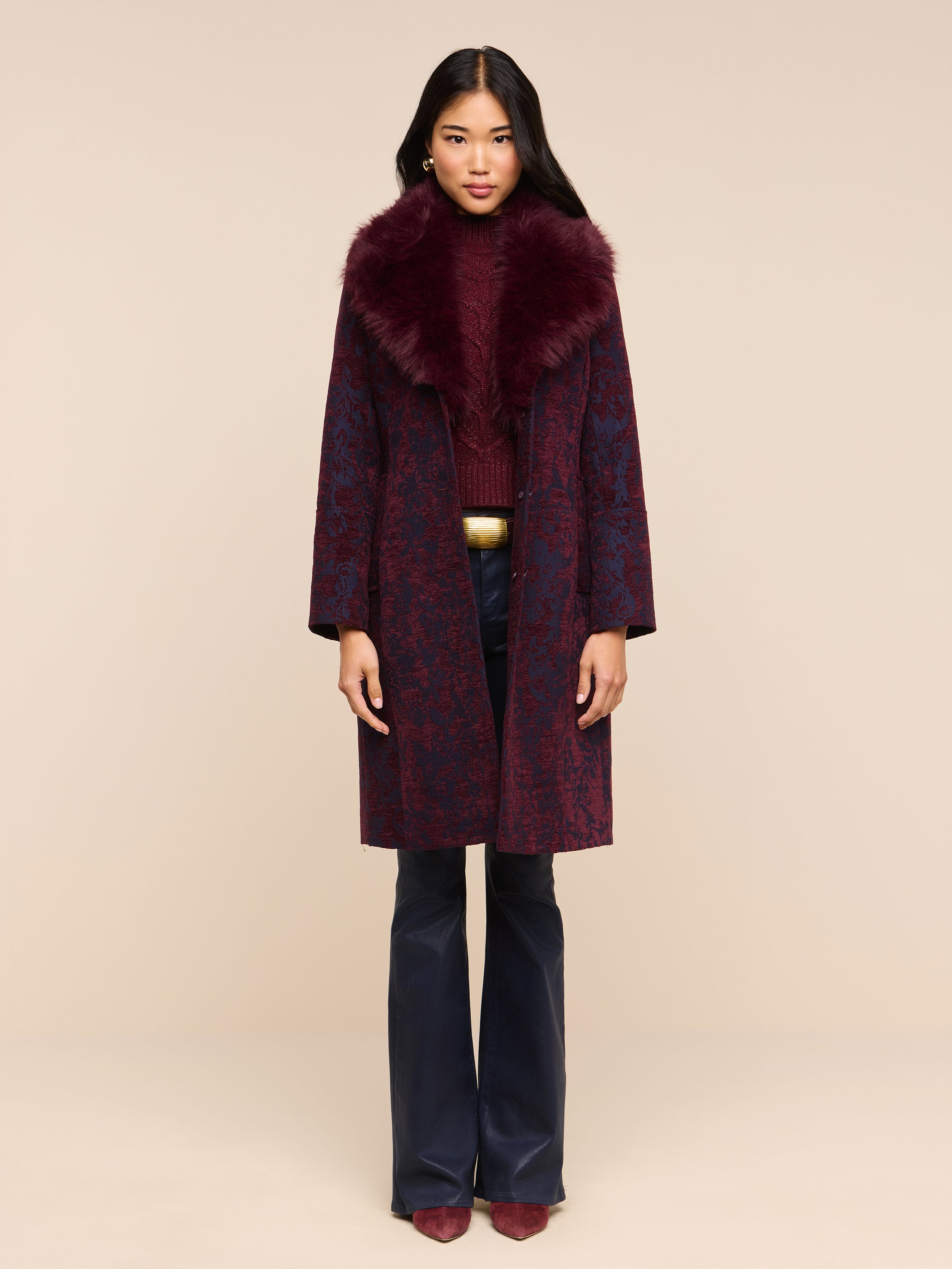 A model wears a long patterned coat with a dark burgundy faux fur collar over a matching sweater and dark Marty Coated Flare Jean.