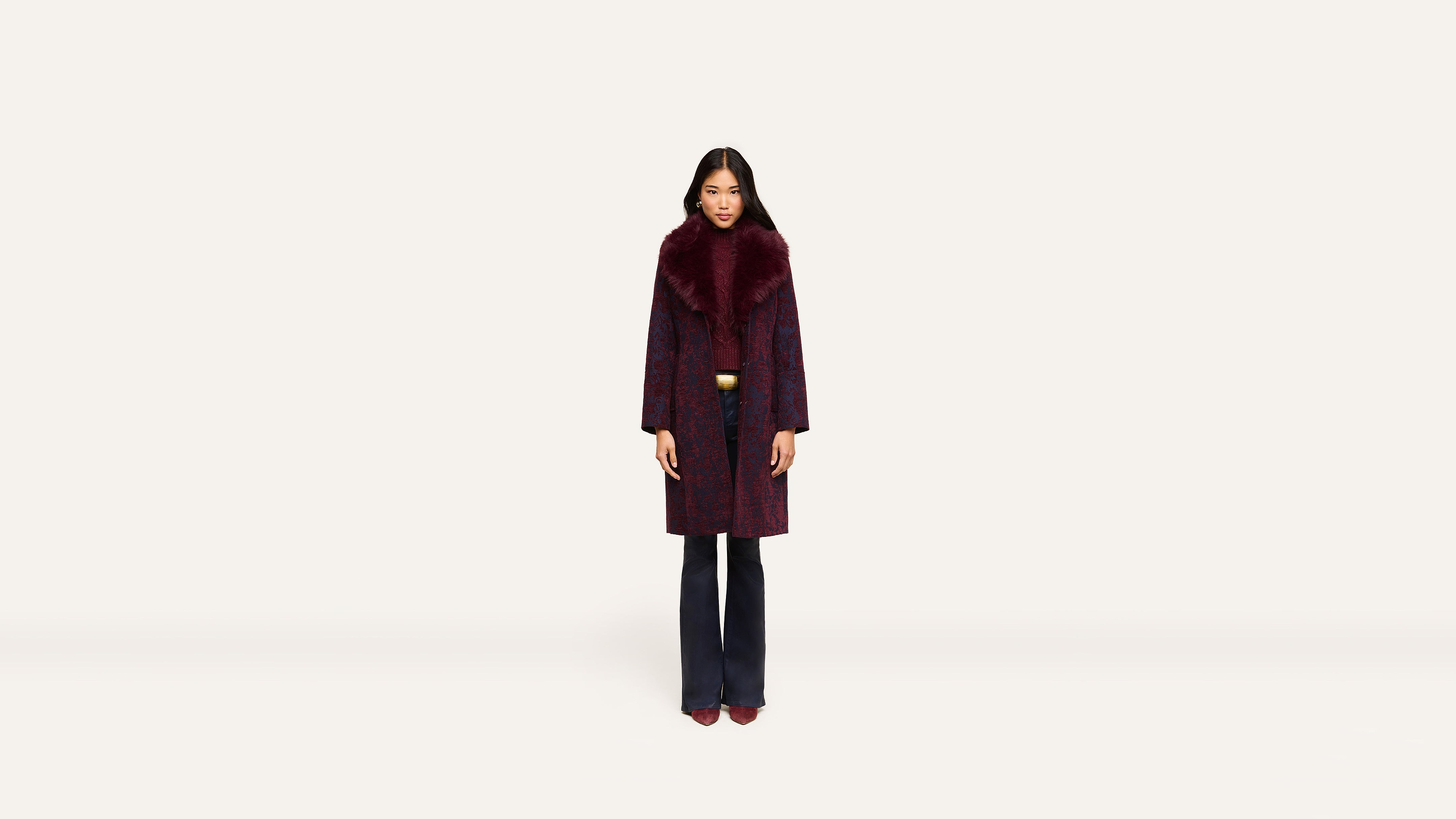 A woman stands facing forward, wearing a long, dark burgundy coat with a wide fur collar and matching burgundy shoes against a plain white background.