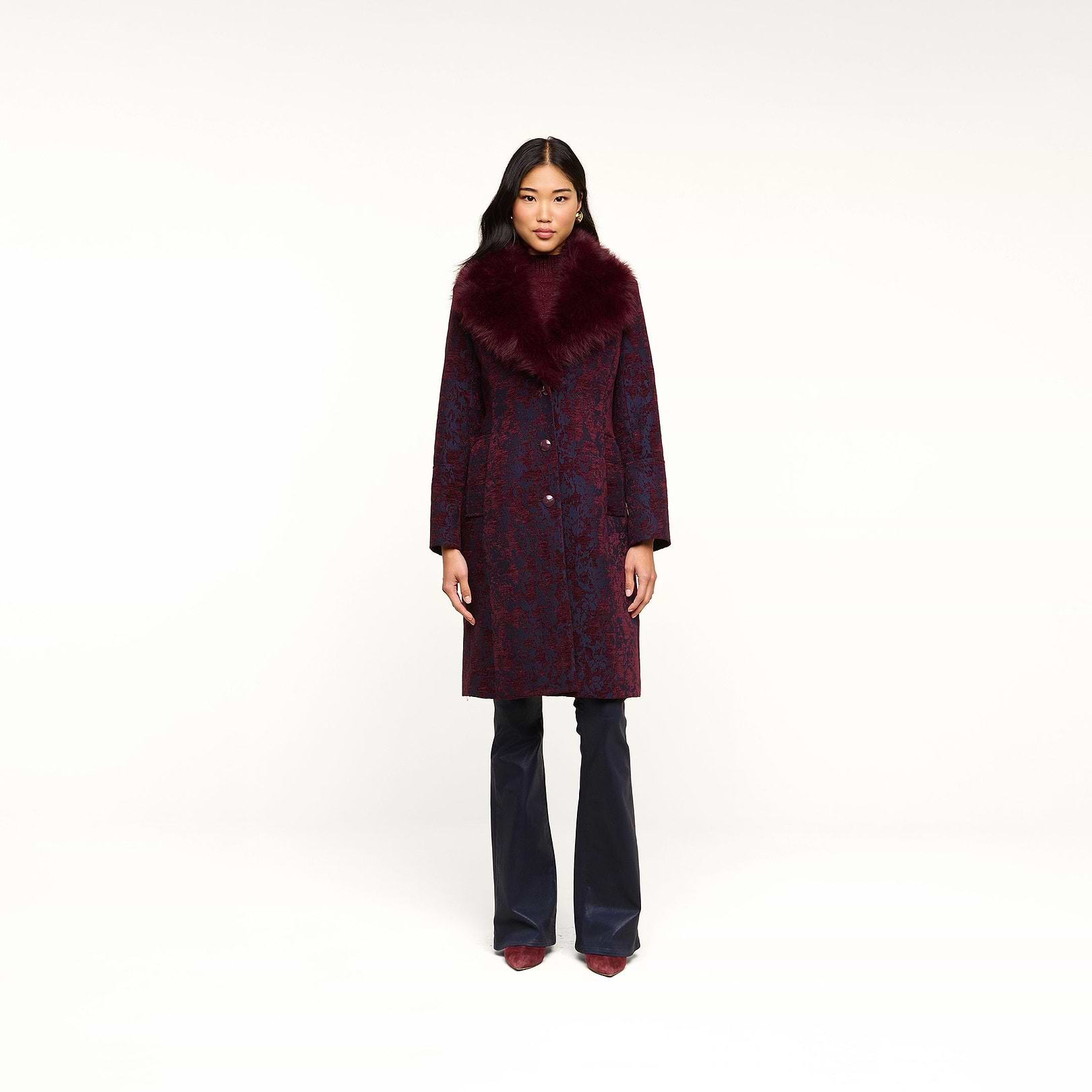 A woman stands facing forward against a plain white background, wearing the Laney Jacquard Coat featuring a vintage-inspired design with a large burgundy faux fur collar, paired with navy pants and burgundy shoes.