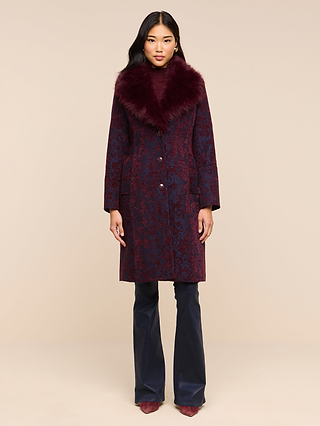 A woman stands against a beige background wearing the Laney Jacquard Coat, which features a vintage-inspired design with a large faux fur collar, styled with navy pants and burgundy shoes.