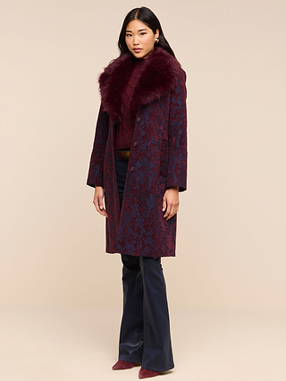A woman stands against a beige background wearing the Laney Jacquard Coat—a burgundy, vintage-inspired outerwear piece with a large faux fur collar—paired with dark pants and burgundy shoes.