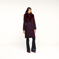 A woman with long, dark hair and a relaxed expression stands against a white background, wearing the Laney Jacquard Coat—a long burgundy coat featuring a large faux fur collar—paired with dark pants and pointed shoes for a vintage-inspired outerwear look.