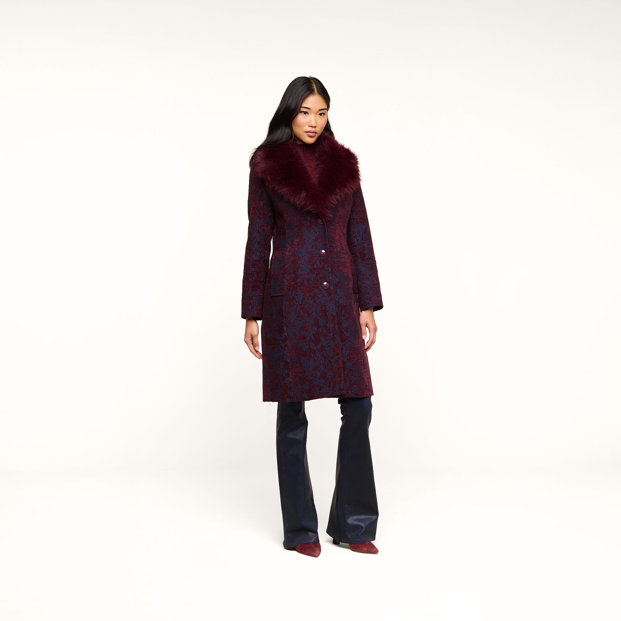 A woman with long, dark hair and a relaxed expression stands against a white background, wearing the Laney Jacquard Coat—a long burgundy coat featuring a large faux fur collar—paired with dark pants and pointed shoes for a vintage-inspired outerwear look.