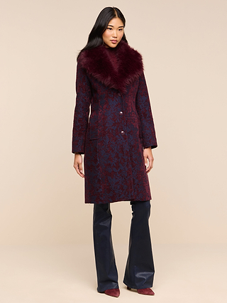A woman wearing the Laney Jacquard Coat—a long, vintage-inspired piece with a large faux fur collar—pairs it with navy flared pants and burgundy shoes as she stands against a plain beige background.