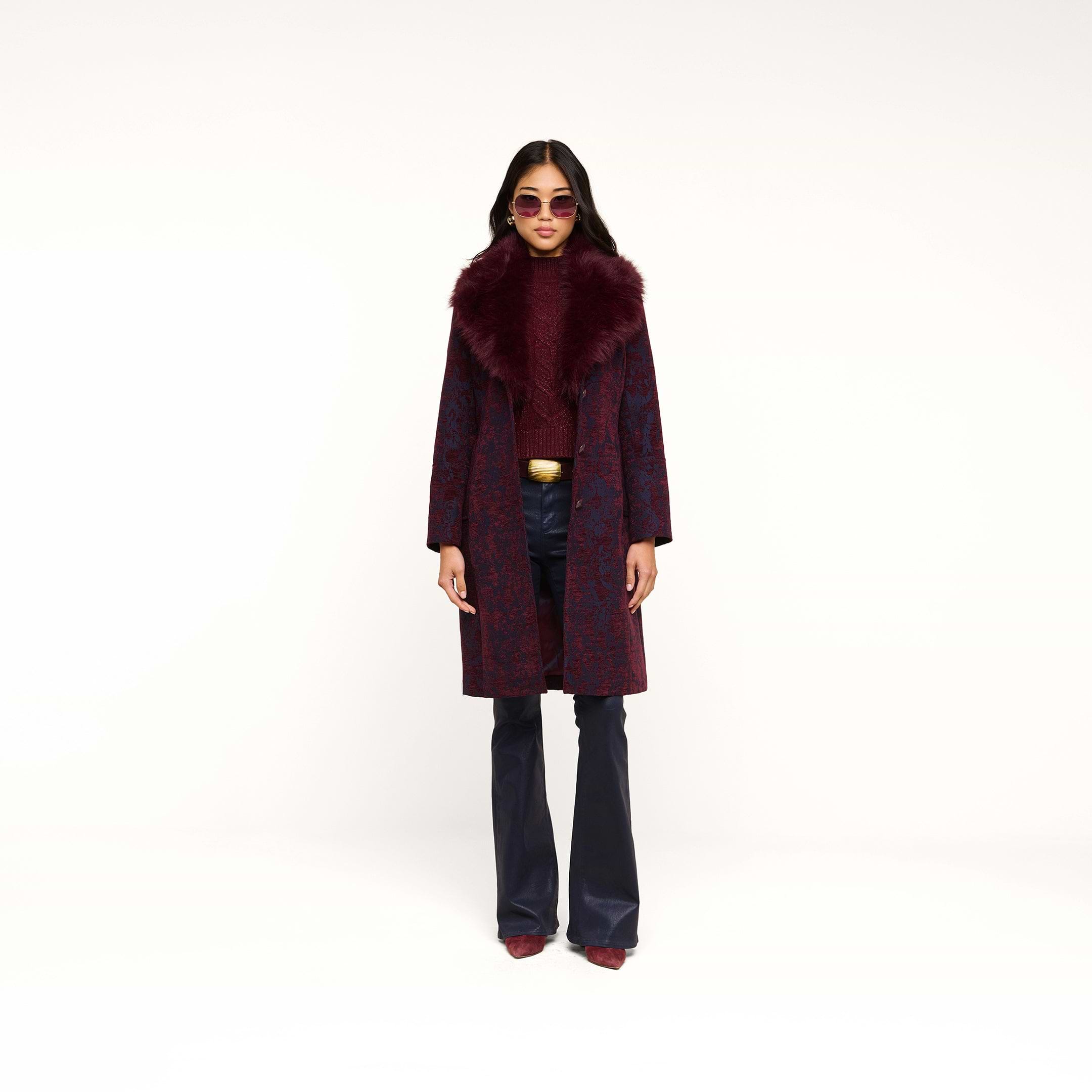 A woman stands against a plain white background wearing the Laney Jacquard Coat—a vintage-inspired burgundy coat with a large faux fur collar—paired with dark flared pants, a maroon top, pointed shoes, sunglasses, and a gold belt.