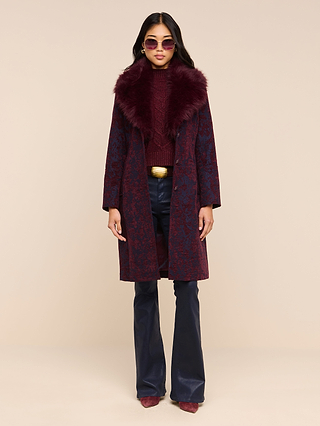 Wearing the Laney Jacquard Coat with its luxurious faux fur collar, a woman stands against a plain beige background. Her vintage-inspired outerwear ensemble is completed by a maroon sweater, navy blue pants, matching shoes, pink sunglasses, and a wide gold belt.