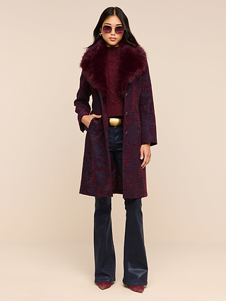 A woman wearing sunglasses, the Laney Jacquard Coat with a faux fur collar, a burgundy sweater, dark flared pants, maroon shoes, and a gold belt stands against a beige background with one hand in her pocket. This vintage-inspired outfit exudes effortless sophistication.