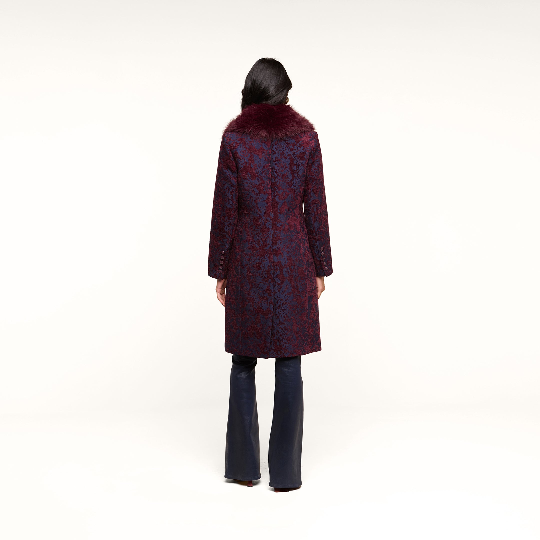 A person with long dark hair, seen from behind, wears the Laney Jacquard Coat—a knee-length, dark red jacquard coat featuring a maroon faux fur collar—paired with dark pants and standing against a plain white background.