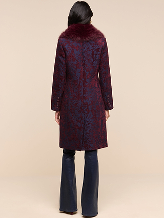 A woman stands with her back to the camera, wearing the Laney Jacquard Coat—a long, dark purple coat with a faux fur collar—paired with navy blue flared pants, against a plain beige background.