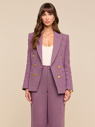 A woman with long brown hair is wearing the Kyomi Double-Breasted Blazer—a tailored purple herringbone piece with gold buttons—over a light top. She stands against a plain beige background, looking directly at the camera with a neutral expression.