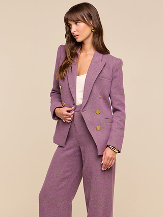 A woman stands against a plain beige background, looking to the side, dressed in the Kyomi Double-Breasted Blazer in mauve with gold buttons, paired with a white top and gold accessories.