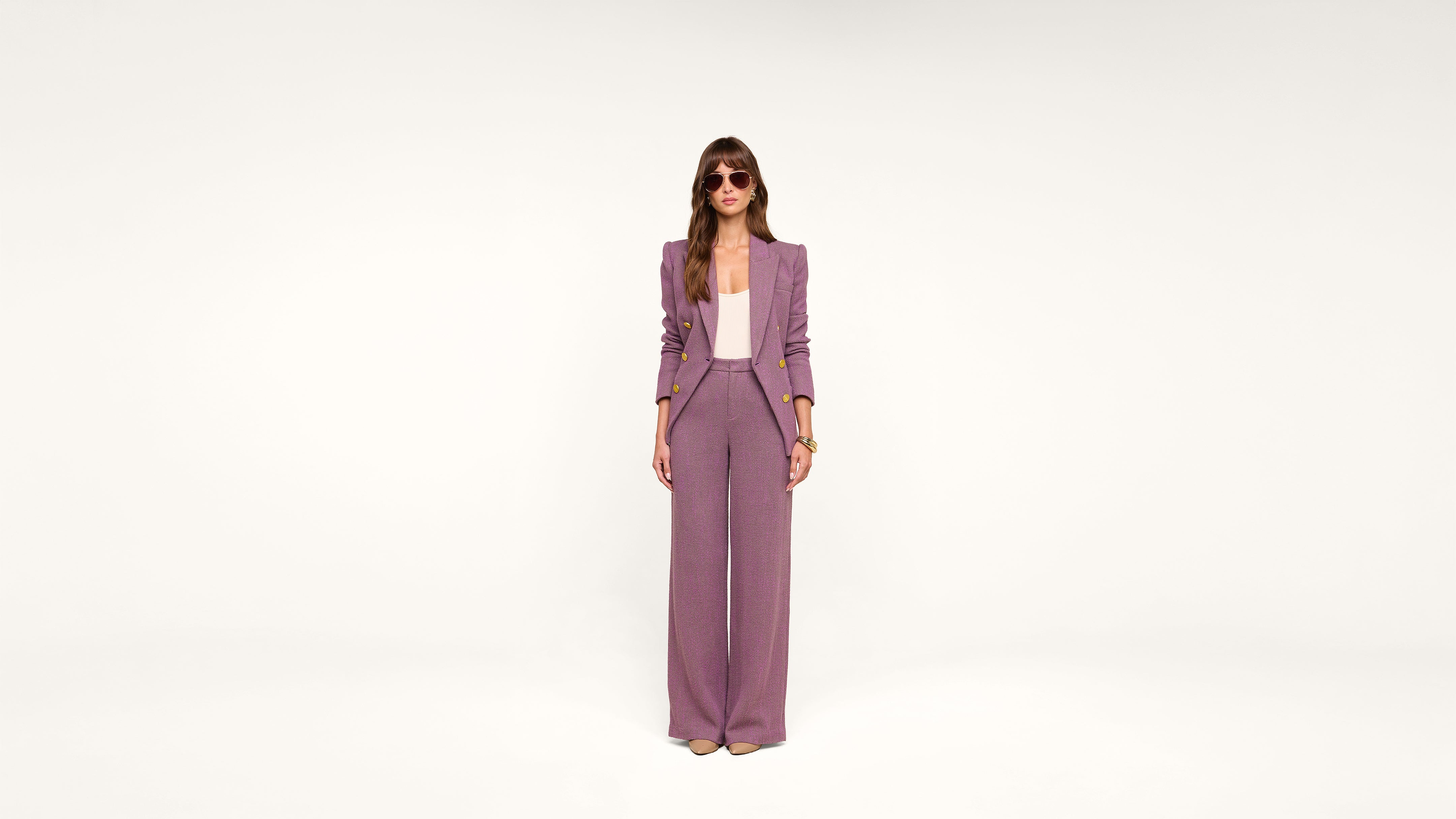 A woman stands in the center, wearing a purple pantsuit, white top, sunglasses, and heels, against a plain white background.