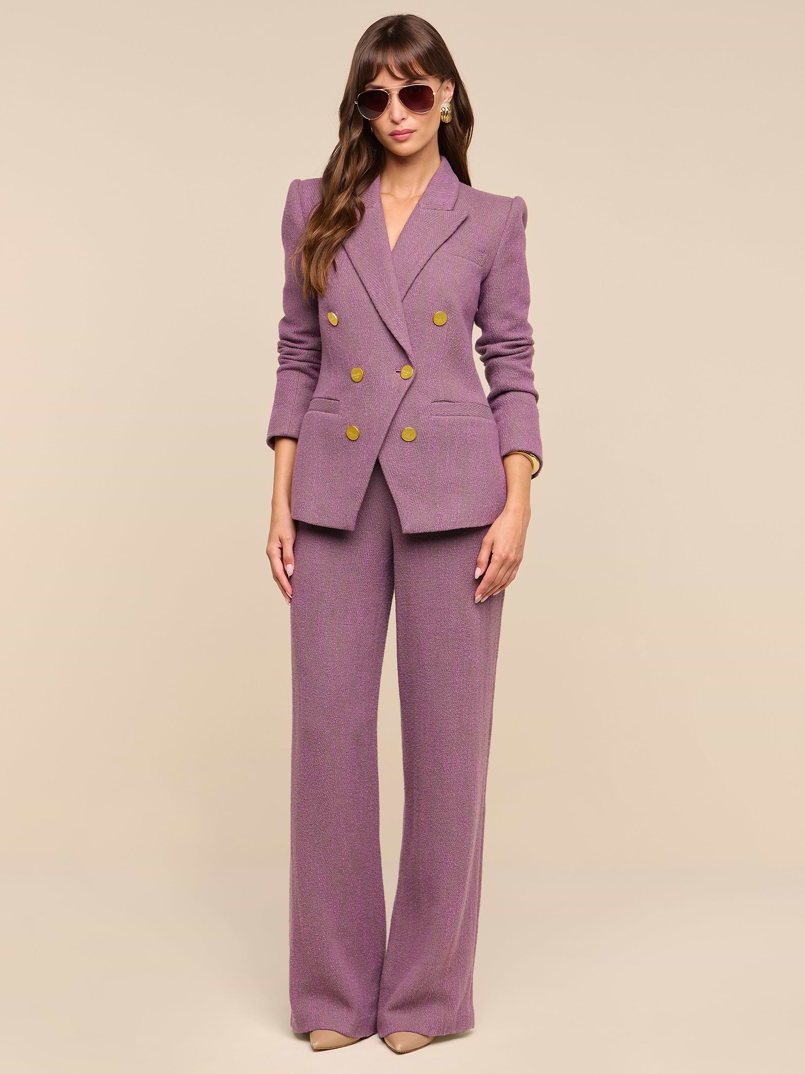A woman stands against a plain white background wearing high-rise Livvy Straight-Leg Trousers in mauve paired with a matching double-breasted blazer, beige heels, and oversized sunglasses. Her hands are relaxed at her sides, and her long hair falls over her shoulders.