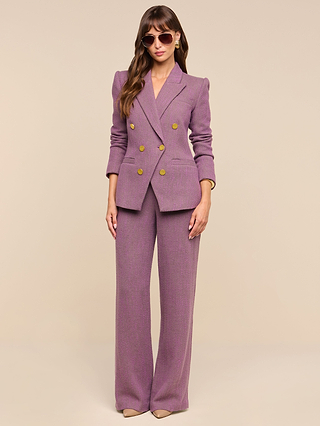 A woman stands against a plain beige background wearing sunglasses and beige heels, styled in the Livvy Straight-Leg Trouser—a mauve high-rise, double-breasted pantsuit with gold buttons, crafted from Italian herringbone weave.