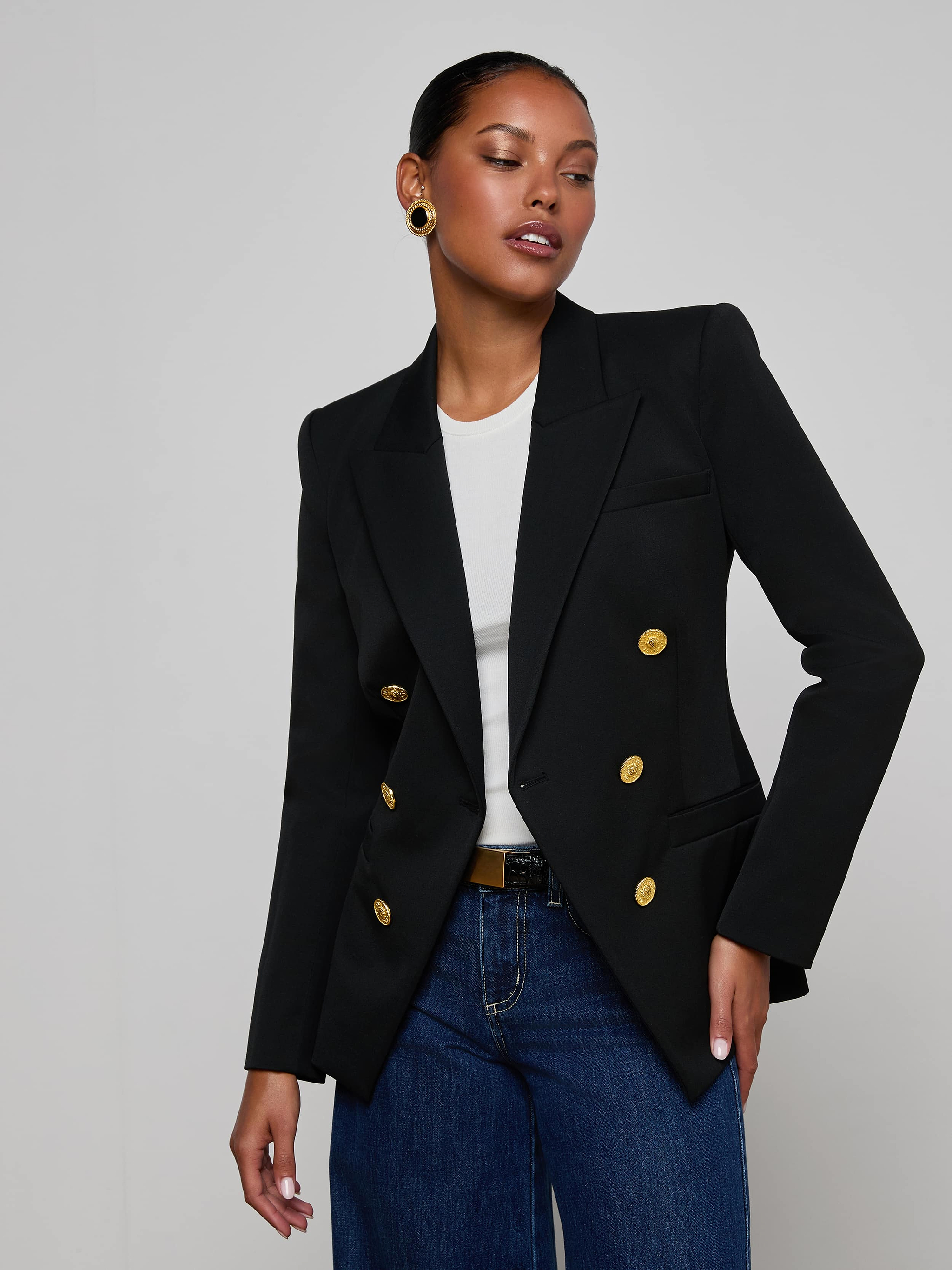 A woman wearing the Kyomi Double-Breasted Blazer with gold buttons, styled over a white shirt and blue jeans, stands against a plain light background.