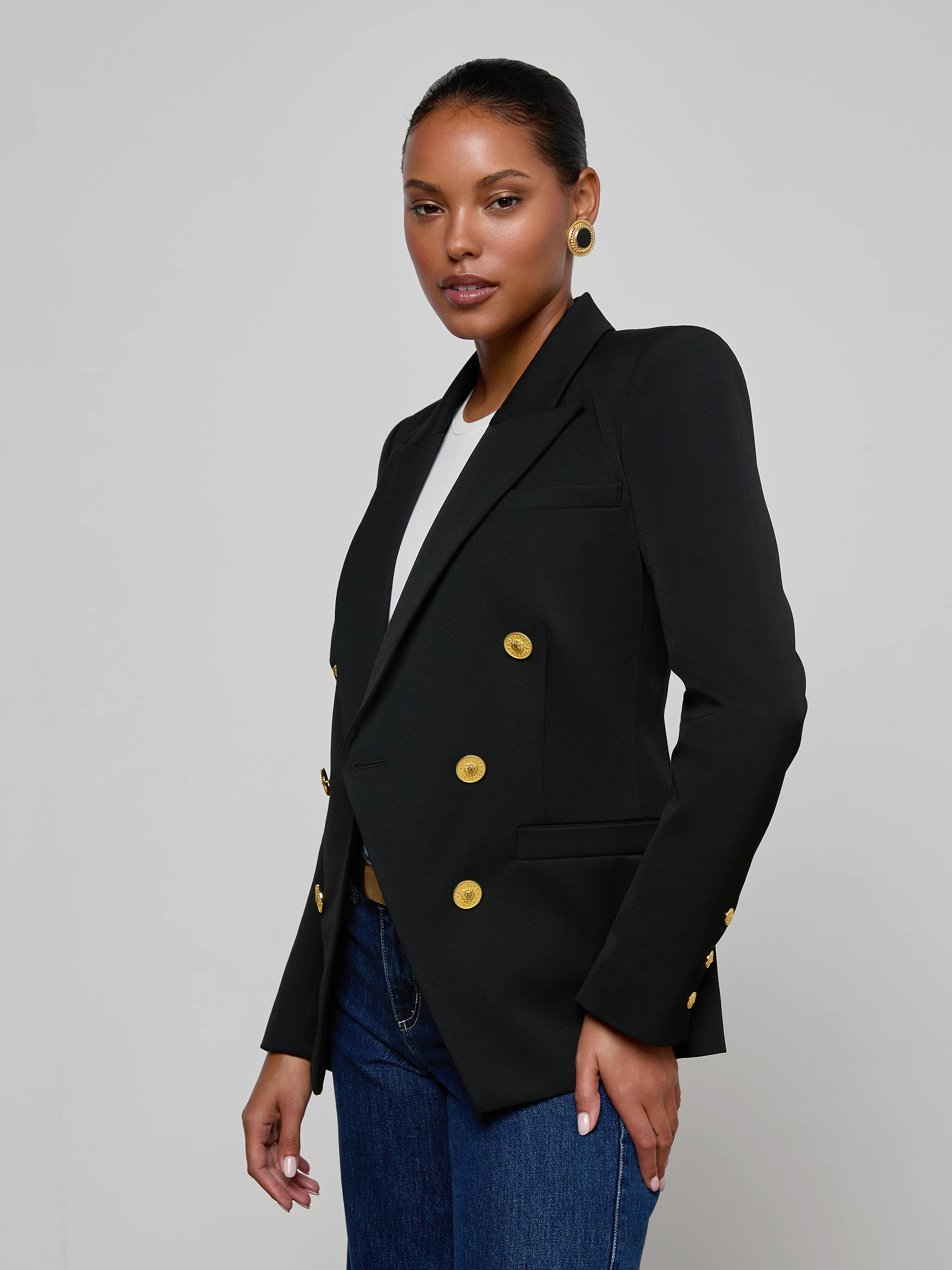 A woman stands against a plain background wearing the Kyomi Double-Breasted Blazer in black with gold buttons, styled over a white top, blue jeans, and accessorized with gold earrings. Her hair is pulled back and she poses with one hand relaxed by her side.