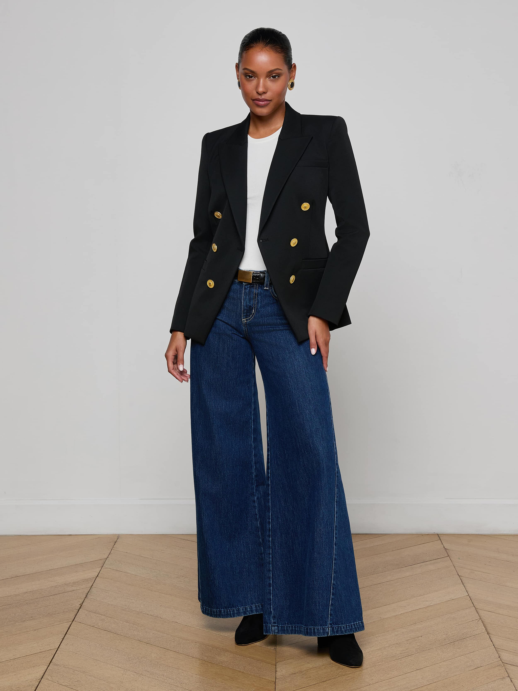 A person stands against a plain background, wearing the Kyomi Double-Breasted Blazer—tailored in black with gold buttons—styled with a white shirt, wide-leg blue jeans, and black shoes.