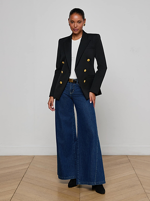 A woman stands indoors, confidently posing in the L'AGENCE Kyomi Double-Breasted Blazer with gold buttons, styled with a white top, wide-leg blue jeans, and black shoes against a light wall and wood floor.